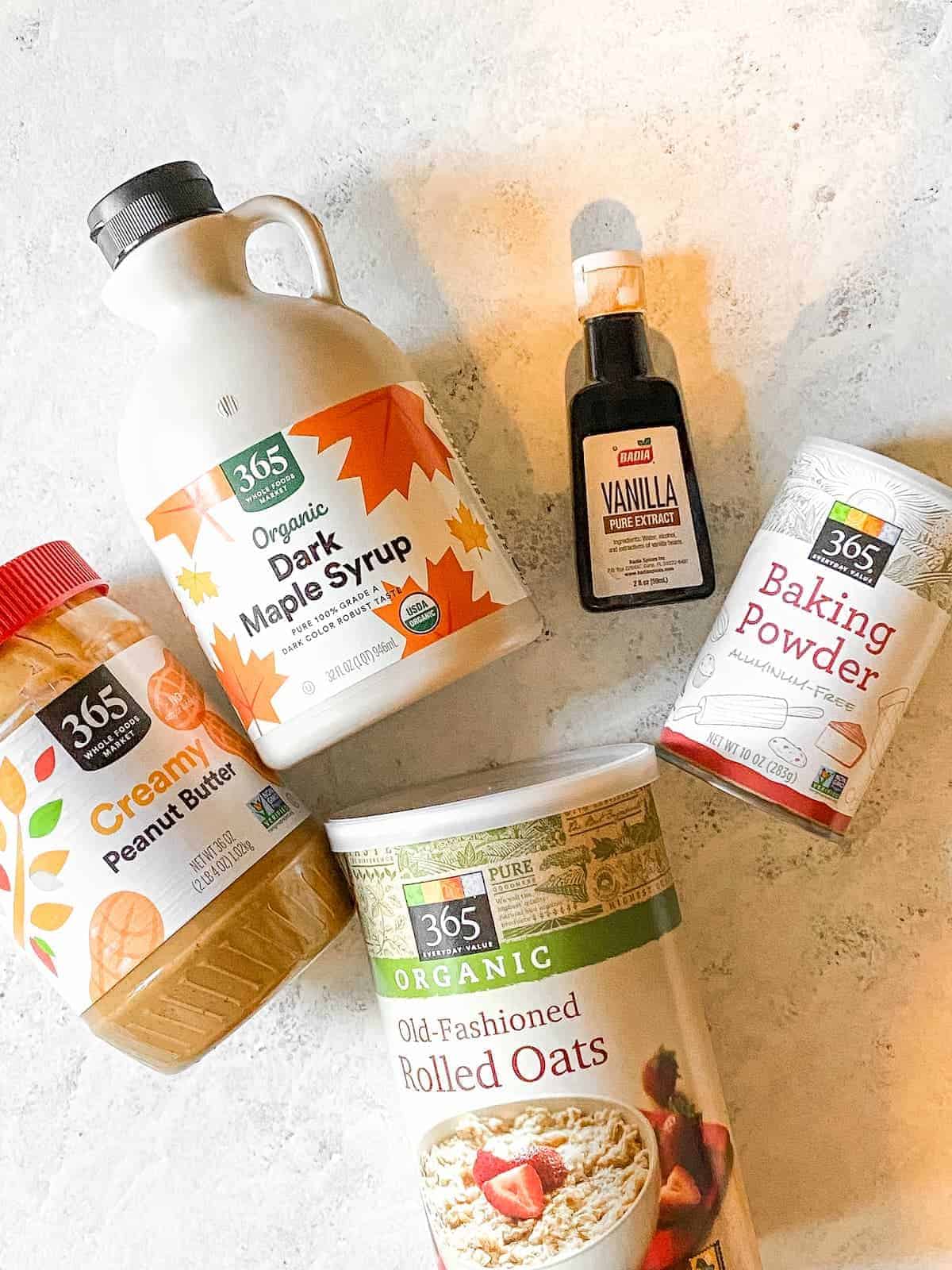 A tub of peanut butter, maple syrup container, vanilla extract, baking powder and rolled oats.