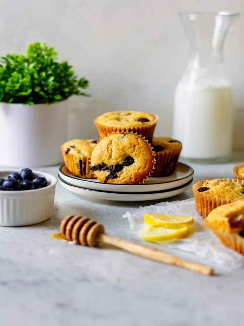 Muffins stacked on a small white plate with a cup of blueberries and a honey dipper in front.