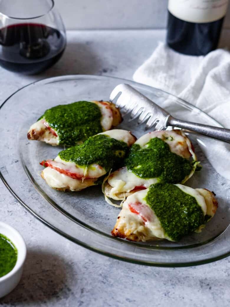 four chicken breasts topped with tomato, mozzarella and pesto on a platter with a pair of tongs and glass of wine in the background