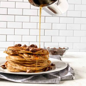 maple syrup being poured onto a stack of banana pancakes