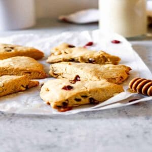 A honey dipper in front of white chocolate and cranberry scones on crinkled wax paper.