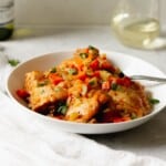 Chicken osso buco in a white bowl in front of a glass of white wine.