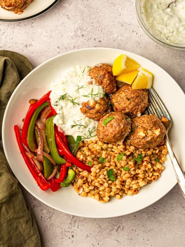 A white plate of Greek meatballs with couscous and red peppers topped with parsley and dill.