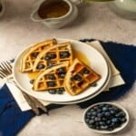 Blueberry waffle on a plate on top of a book and blue napkin with silver forks and a syrup pourer.