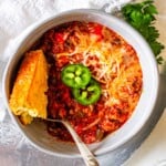 A bowl of chili topped with jalapeno slices and a piece of cornbread.