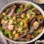 A bowl of Caesar salad with steak and croutons.