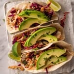 Three tacos topped with slaw and avocado garnished with lime wedges.