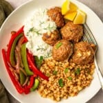 A white plate of Greek meatballs with couscous and red peppers topped with parsley and dill.