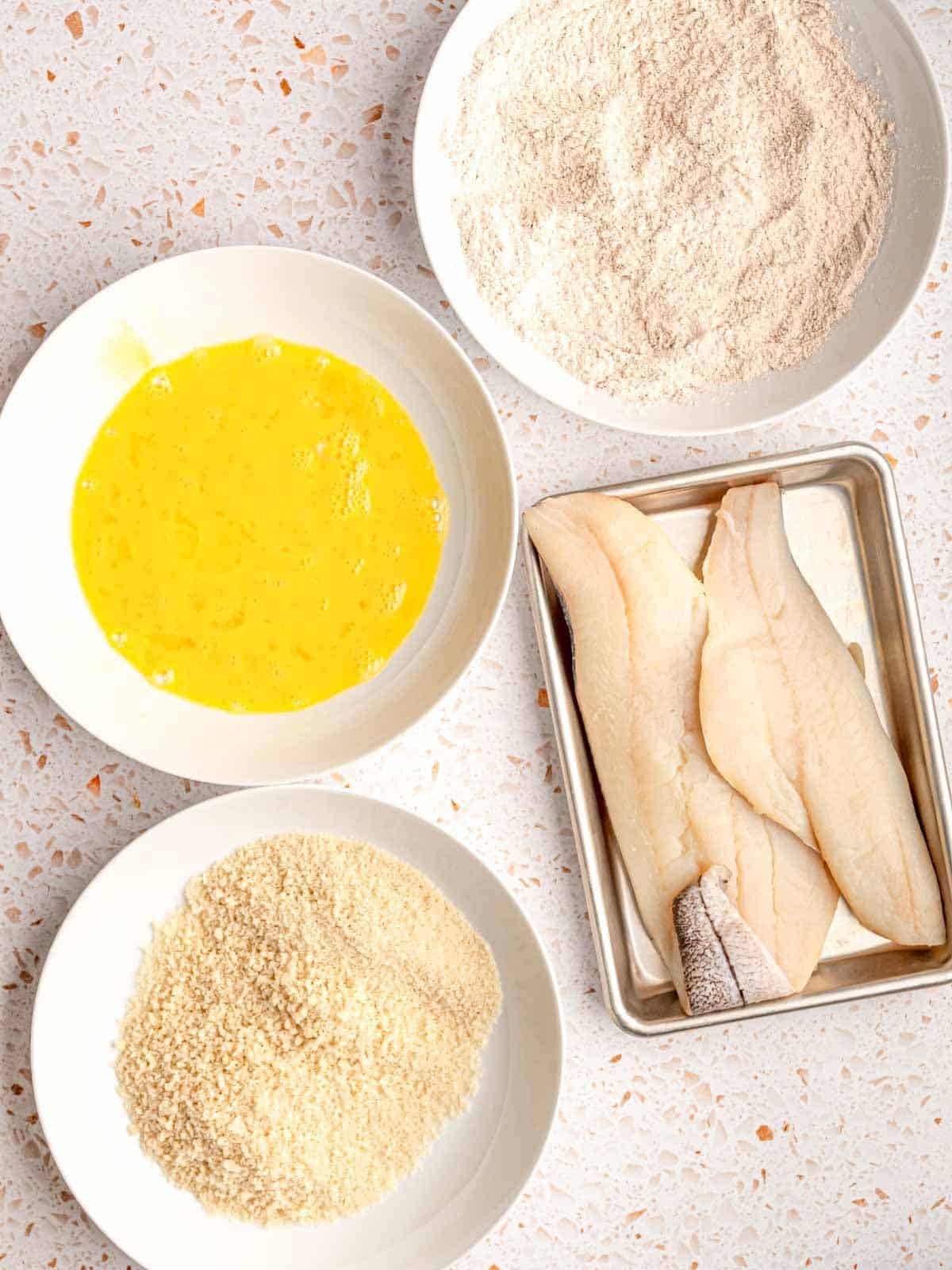 Fish on a metal tray next to bowls of flour, eggs and breadcrumbs.