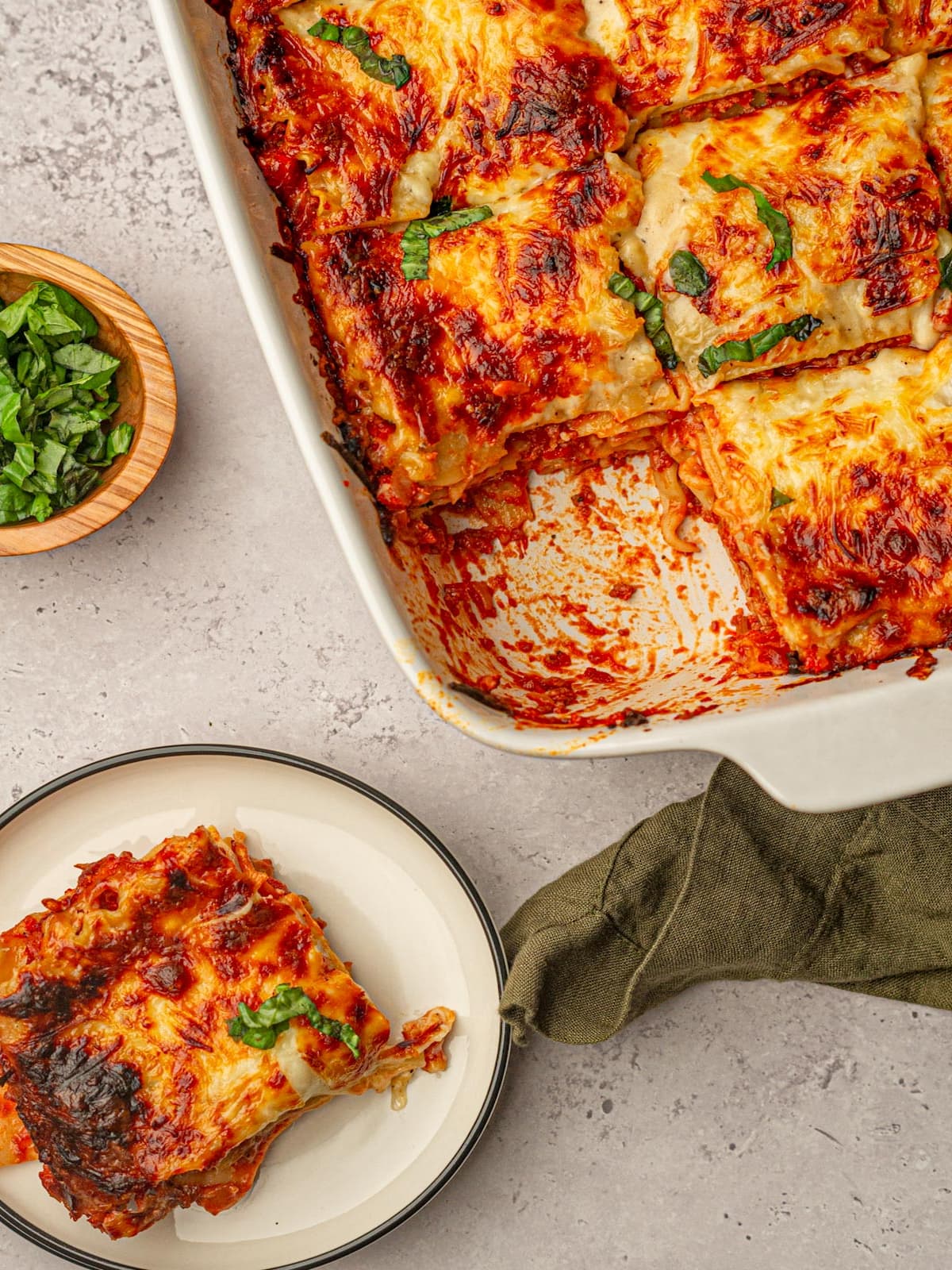A serving of lasagna with béchamel sauce on a plate, next to a half-empty baking dish with sliced portions and a bowl of fresh basil.