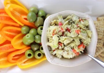 Chopped artichoke hearts, diced red bell peppers, capers and chopped leaks dress ing a white bowl surrounded by chopped vegetables.