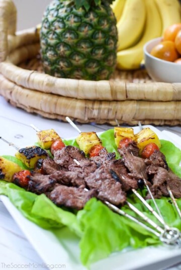 Steak skewers with pineapple and cherry tomatoes on a white dish laid on lettuce.