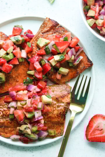 Grilled salmon on a white plate topped with strawberry, red onion and avocado salsa.