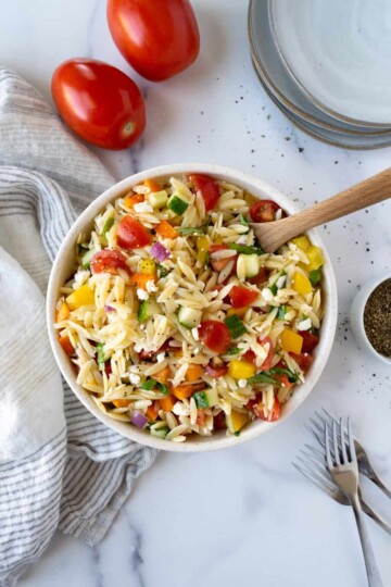 Orzo with tomatoes, bell peppers, cucumbers, red onion, feta cheese in a white bowl.