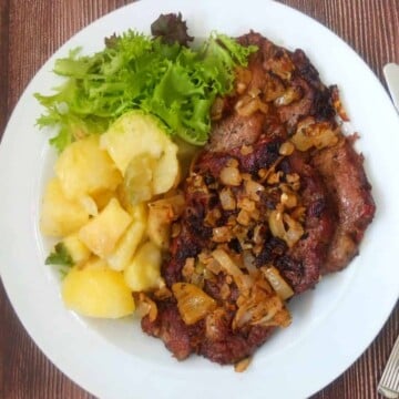 Grilled pork steak covered with onions, on a white plate with potatoes and lettuce.