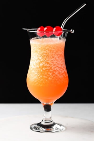 Frozen orange drink in a tall glass with 3 cherries on a cocktail skewer.