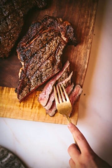 Hand holding a fork resting on cut pieces of grilled ribeye steak resting on a wooden board.
