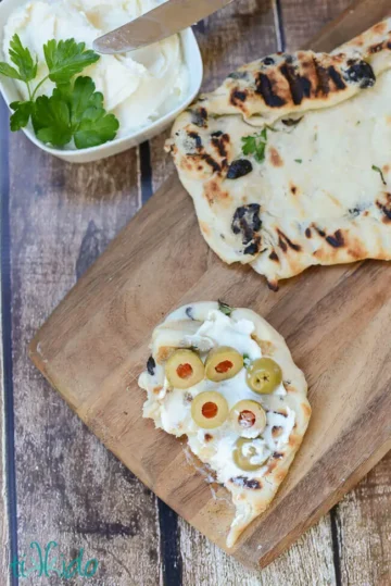 Grilled flatbread on a wooden board with whipped feta and green olives.