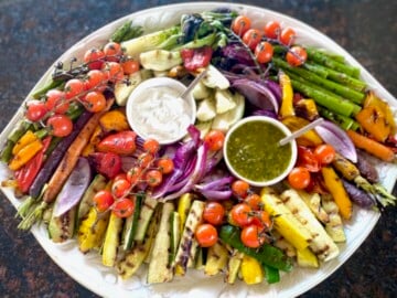 Rainbow grilled vegetables on a white platter with white and green dip.