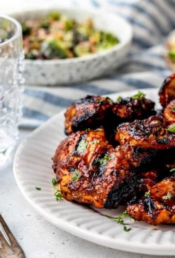 Marinated sticky bbq grilled chicken thighs stacked on a white plate, garnished with parsley.