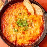 Skillet filled with creamy chicken enchilada dip topped with melted cheese, garnished with chopped scallions, and surrounded by tortilla chips.