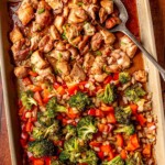 Close-up of sesame chicken garnished with green onions alongside vibrant roasted vegetables on a sheet pan, ready to serve.