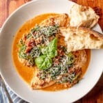 A plated serving of Tuscan chicken topped with spinach and sun-dried tomatoes in a creamy sauce, garnished with fresh basil and shredded Parmesan cheese. Served alongside crusty bread for dipping.
