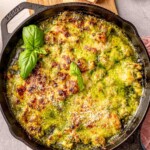 A close-up of baked chicken pesto gnocchi in a cast-iron skillet, topped with golden melted cheese and fresh basil leaves.