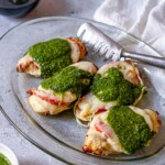 Close-up of air fryer Caprese chicken on a glass platter, showing melted cheese, juicy tomatoes, and vibrant basil pesto with a metal serving tong.