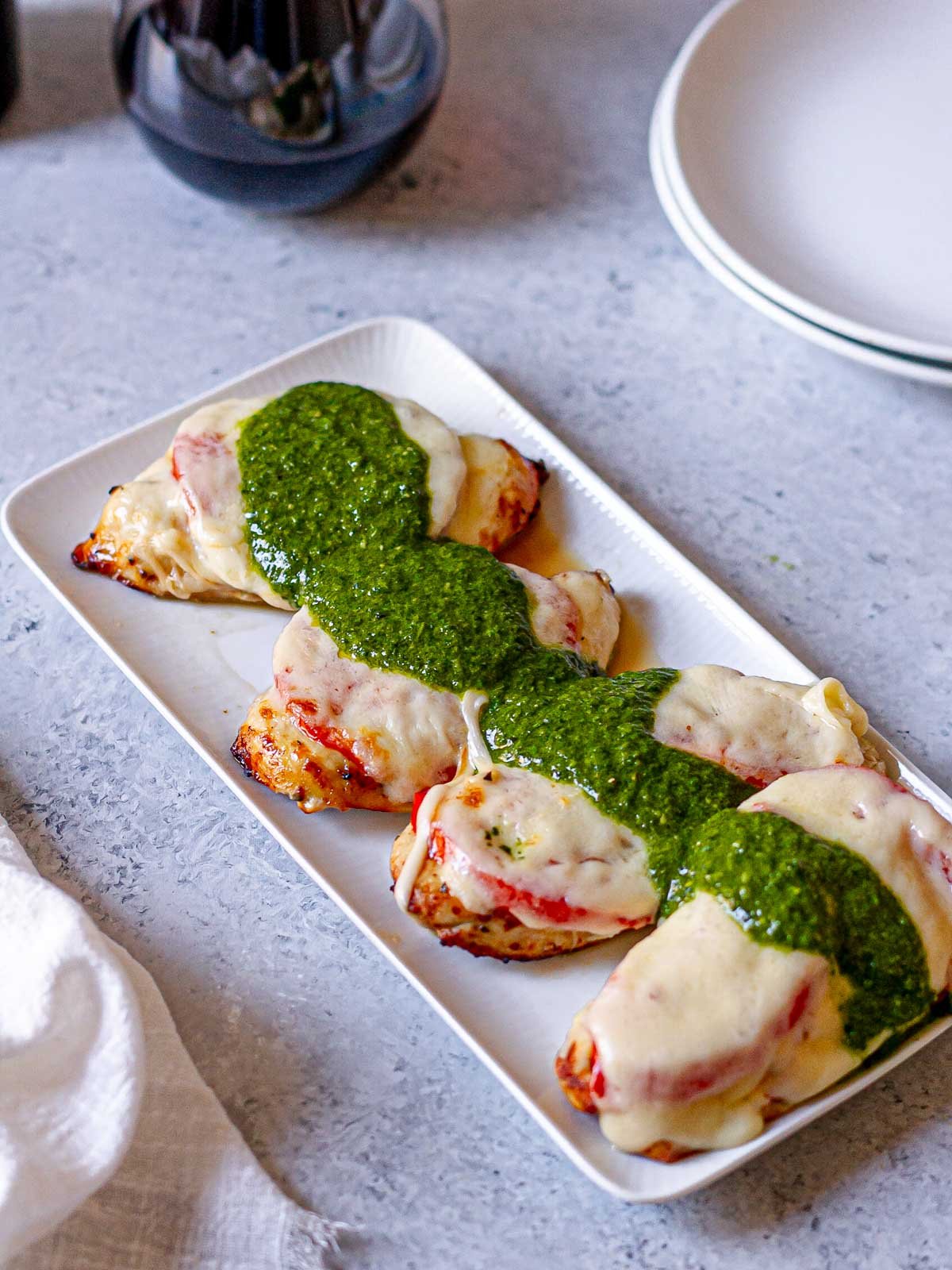 Four baked chicken breasts topped with sliced tomato, melted mozzarella, and bright green pesto on a rectangular white platter with red wine in the background.