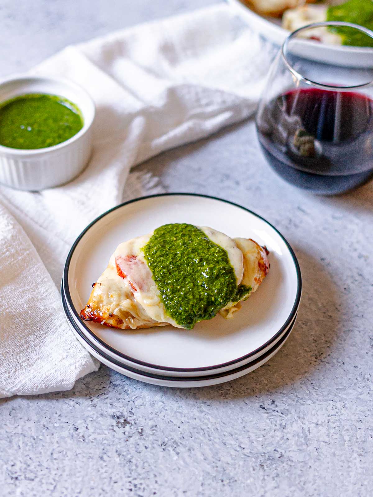 Single serving of Caprese chicken with tomato, melted mozzarella, and pesto on a small white plate beside a ramekin of pesto and a glass of red wine.