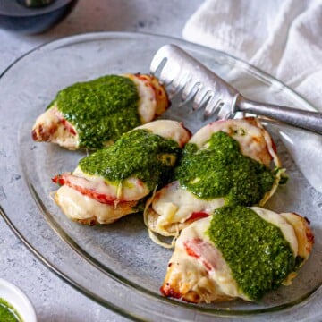 Close-up of air fryer Caprese chicken on a glass platter, showing melted cheese, juicy tomatoes, and vibrant basil pesto with a metal serving tong.