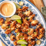 Bang bang chicken skewers with a bowl of dipping sauce, lime wedges, sesame seeds, and scallions on a floral platter.