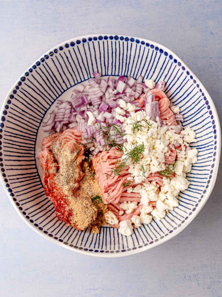 Large bowl with ground turkey, diced red onion, feta cheese, dill, and seasonings ready to be mixed.