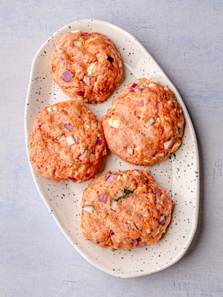 Four uncooked turkey burger patties with visible chunks of red onion and feta on a white speckled platter.