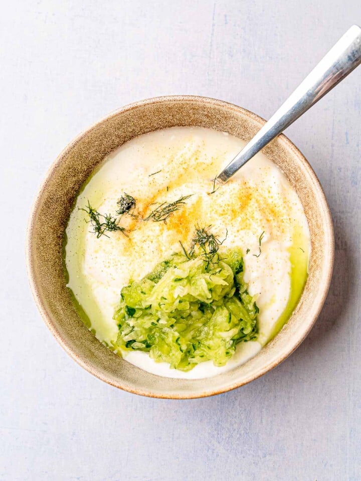 Bowl of yogurt topped with shredded cucumber, dill, and spices ready to be mixed for tzatziki sauce.