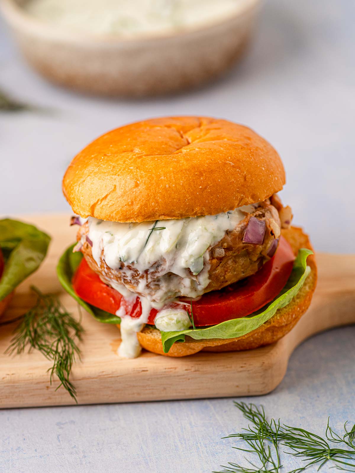 Side view of assembled Greek turkey burger on a wooden board with lettuce and tomato.