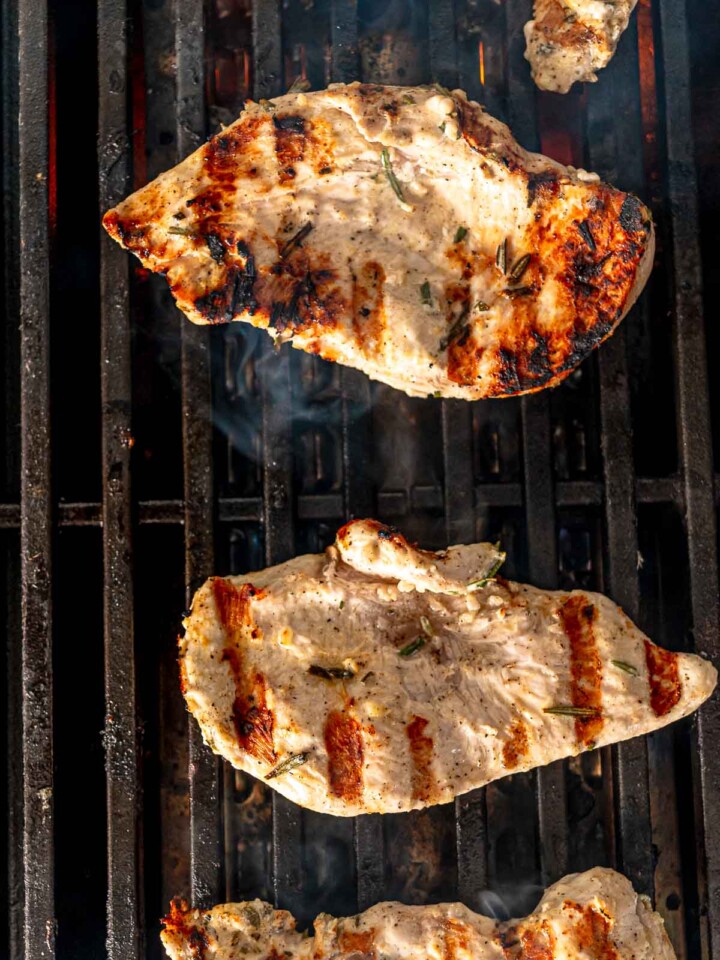 Seasoned chicken breasts cooking on a grill with visible char marks and smoke rising.