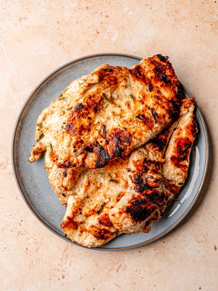 Grilled chicken breasts stacked on a gray plate, showing golden-brown grill marks and bits of rosemary.