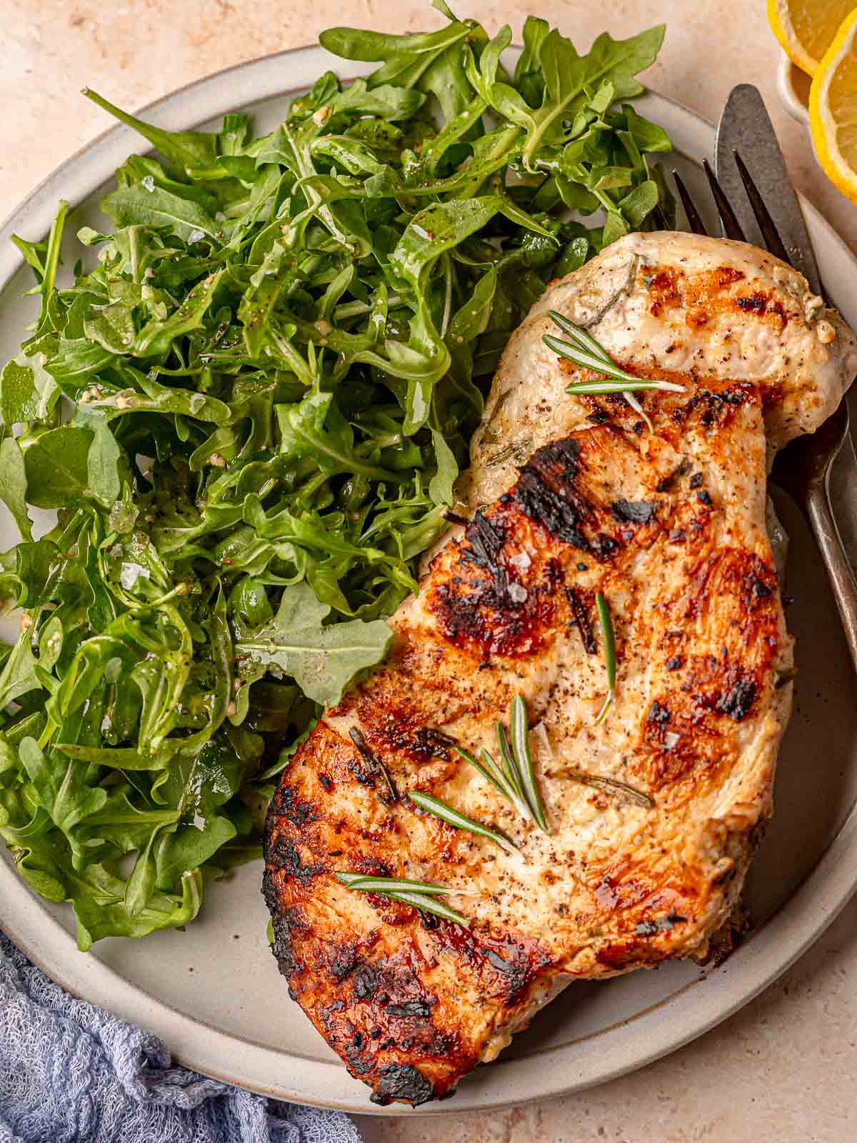 Plate of grilled chicken breast next to lemon slices and fresh arugula salad, ready to serve.