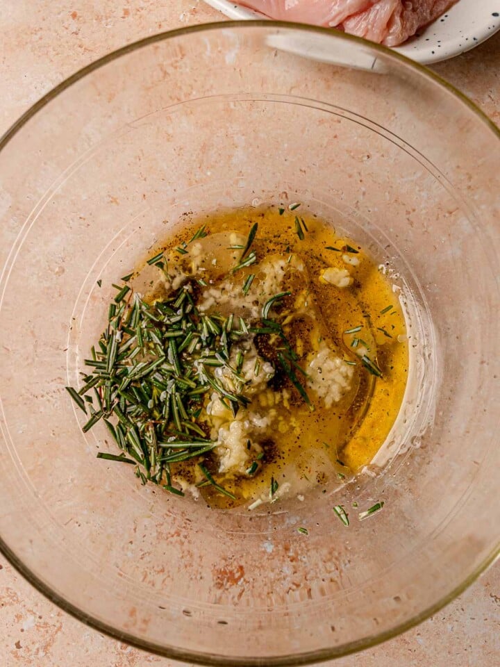 Olive oil, garlic, and chopped rosemary mixed in a glass bowl to marinate chicken breasts.