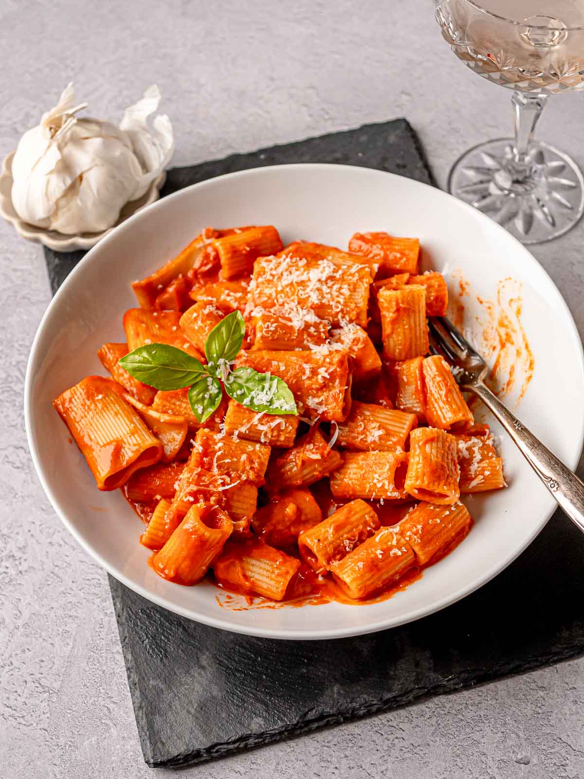 Creamy pink sauce rigatoni topped with fresh basil and cheese, plated on a white bowl with a glass of wine and garlic bulb nearby.