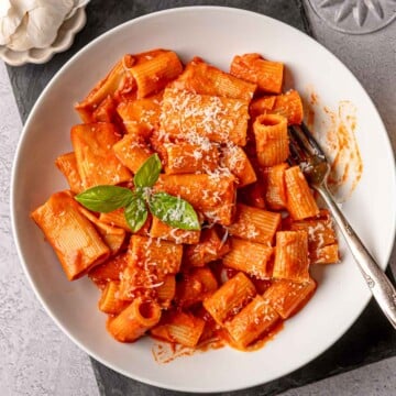Close view of creamy rigatoni pasta in pink sauce, sprinkled with Parmesan and topped with fresh basil leaves, with a fork in the bowl.