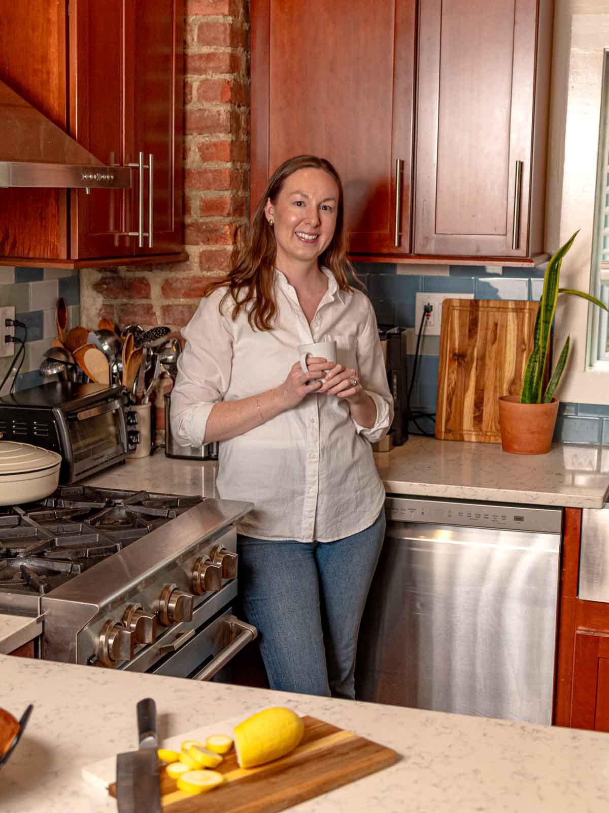 Woman in a white button-up and jeans smiling while holding a white mug and leaning against a kitchen counter with a tiled backsplash and exposed brick wall.