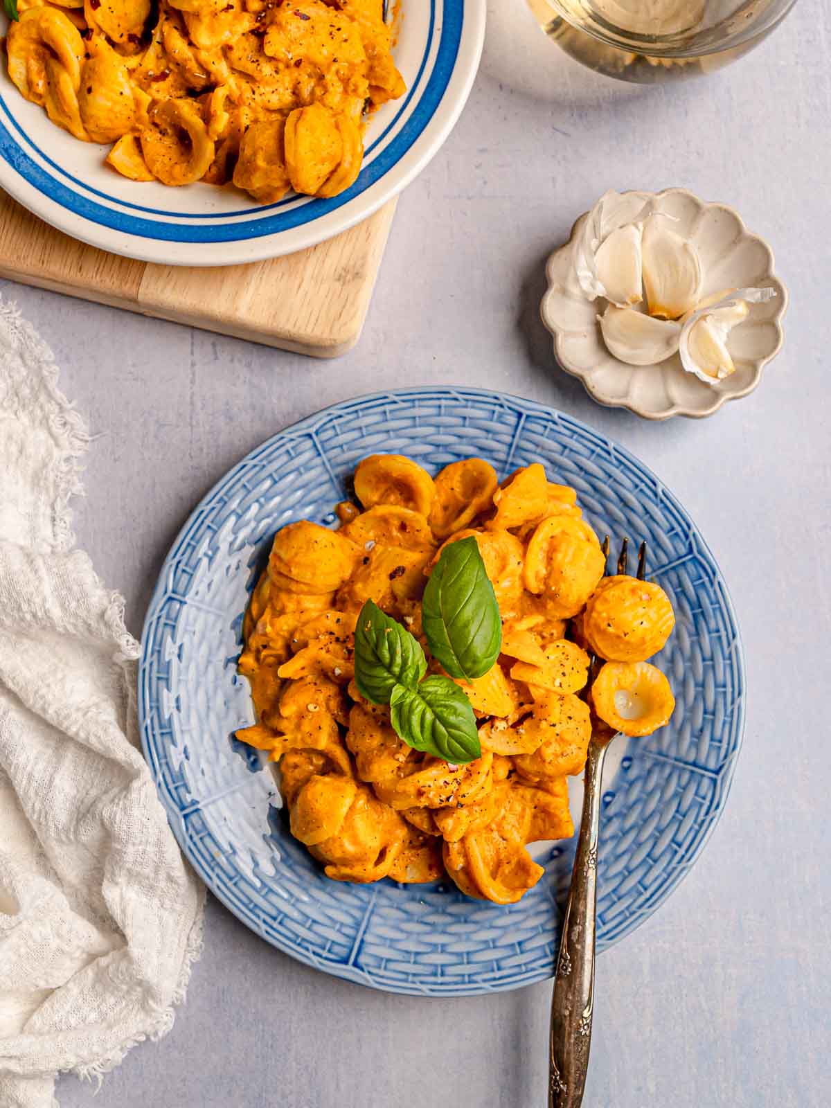 Two servings of vegan pumpkin pasta with basil, styled with garlic cloves and a glass of wine.
