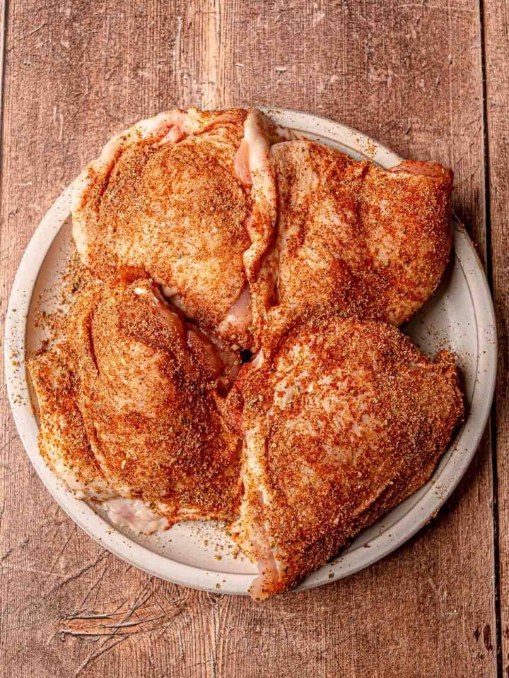 Uncooked chicken thighs coated generously in a red-brown spice rub, arranged on a plate against a rustic wood background.