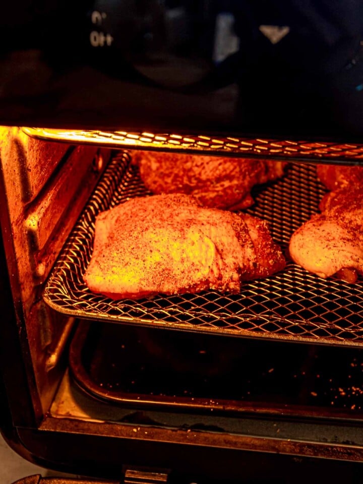 Seasoned chicken thighs cooking inside an air fryer basket under the heating element.