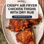 Plate of golden-brown, crispy chicken thighs with a rich dry rub seasoning, garnished with green onions, styled on a speckled platter with text overlay for the recipe.