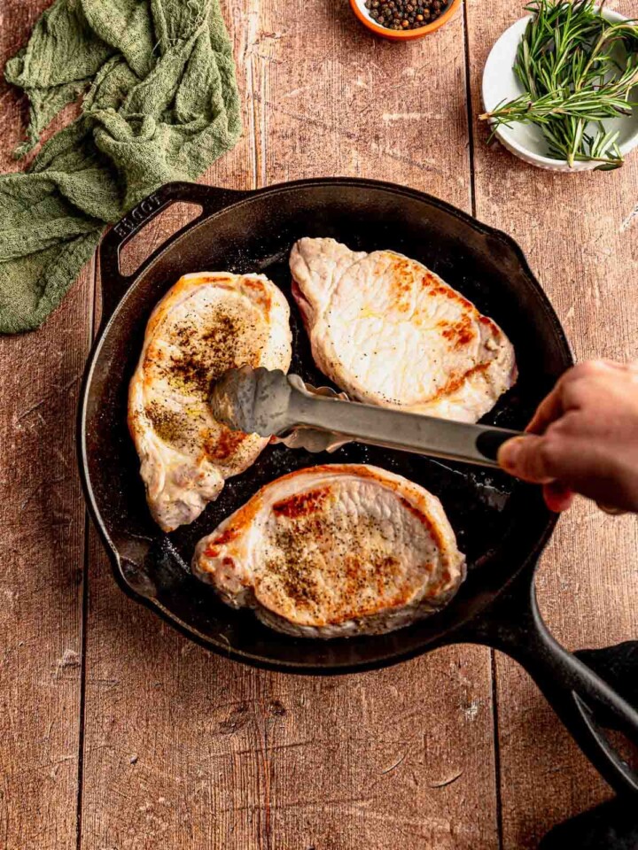 Three pork chops seasoned with salt and pepper searing to golden brown in a black cast iron pan.