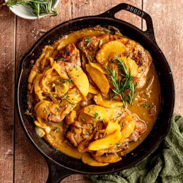 Cooked pork chops nestled in a skillet with caramelized apples, onions, cider sauce, and fresh rosemary.